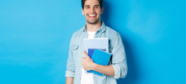 Smiling man studying, holding notebooks and looking happy, standing over blue background.